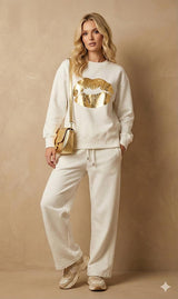 Woman wearing a white sweatshirt with gold design and matching pants, holding a gold handbag against a beige background.