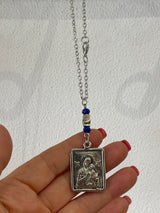 Hand holding a silver car charm with a rectangular icon  of the Virgin Mary and baby Jesus against a white background