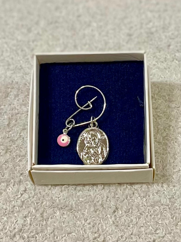 A silver pin with a pink mati charm and a panagia pendant displayed in a box with a blue background.