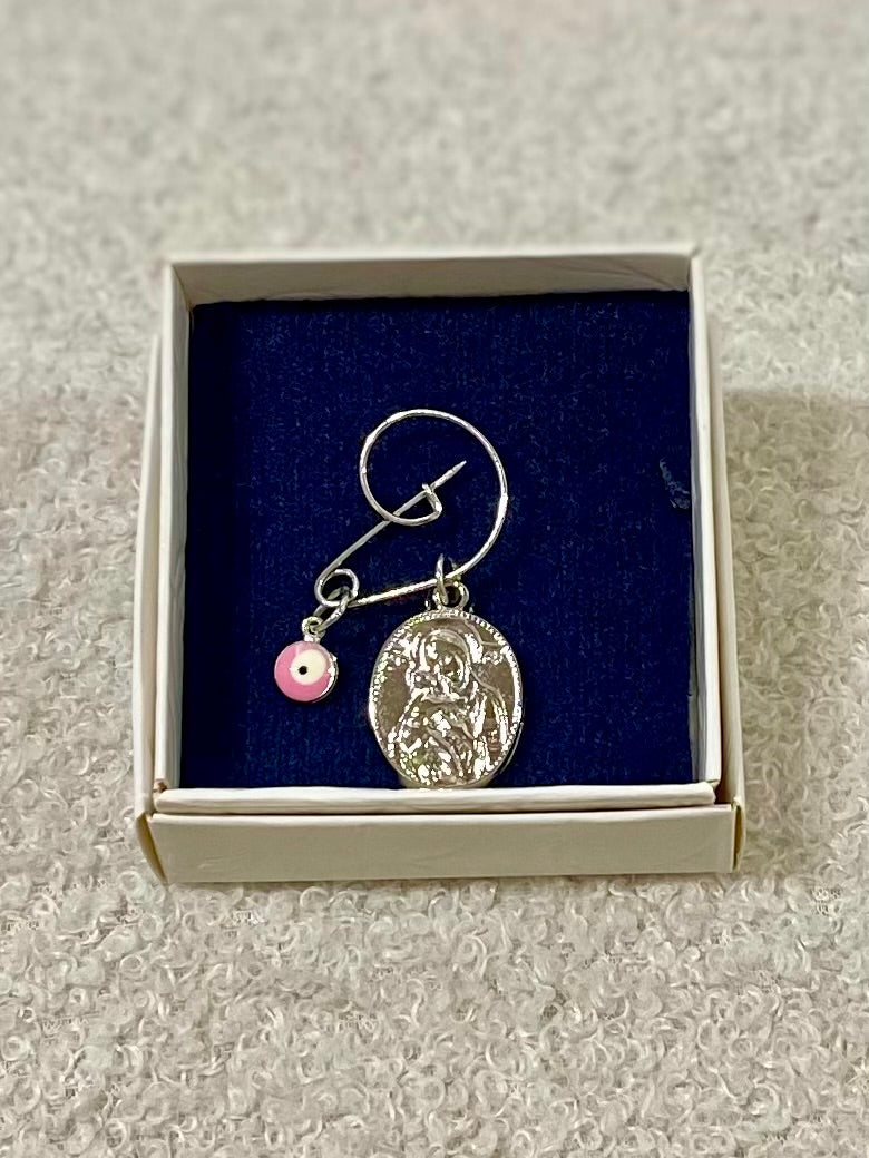 A silver pin with a pink mati charm and a panagia pendant displayed in a box with a blue background.